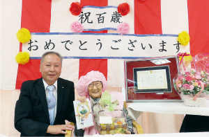「祝百歳おめでとうございます」と掲げられた紅白幕の前で桃色のちゃんちゃんこを着て座っている百歳を迎えた女性と、隣に座った男性の記念写真