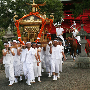 赤い神社のある境内で豪奢な神輿を担いで歩く男性達の写真