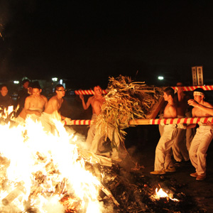 燃え盛る炎の近くで上半身裸に白いズボンを履いた男性数人が、紅白の棒で作られた籠から枯れた葉を炎に焚べている写真