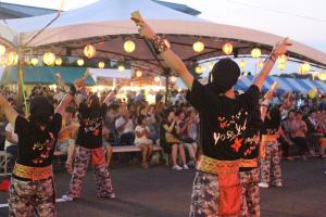 夕暮れの夏祭り会場で、踊りのパフォーマンスを行う女性達とそれを見物する人々の写真