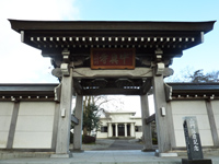 屋根が黒に塗装された寺の門とその近くに石碑が立てられている写真