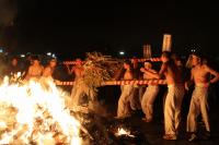 燃え盛る炎の近くで上半身裸に白いズボンを履いた男性数人が、紅白の棒で作られた籠から枯れた葉を炎に焚べている写真