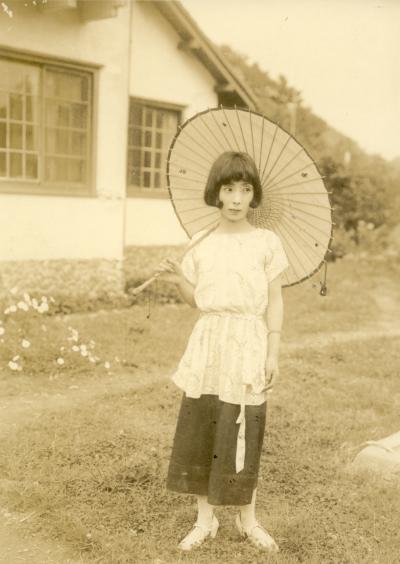 草むらに和傘を差し視線を逸らした白いトップスに黒いボトムスのショートカットの女性が立ち、奥にガラス窓のある家がセピア色にまとめられた写真