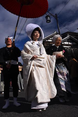 黒い着物姿の年配の女性に手を握られて歩く白無垢姿の女性と傘を差し掛ける男性の写真