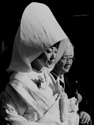 眼鏡をかけている黒い着物を着た年配の女性に手を握られる白無垢姿の女性の写真