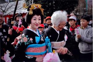 人々が見守る中、黒い着物を着た年配の女性に手を握られて歩く日本髪で華やかな着物姿の女性の写真