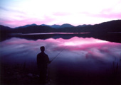 ピンクと紫が混ざりあった空の色を反射している湖のほとりで釣りをしている人の写真