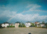 青い空の下に並ぶ複数の家の上に大きな虹がかかっている写真