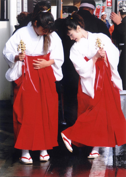 赤と白の巫女服を着て巫女鈴を持った二人の女性が草履の裏を気にしている写真