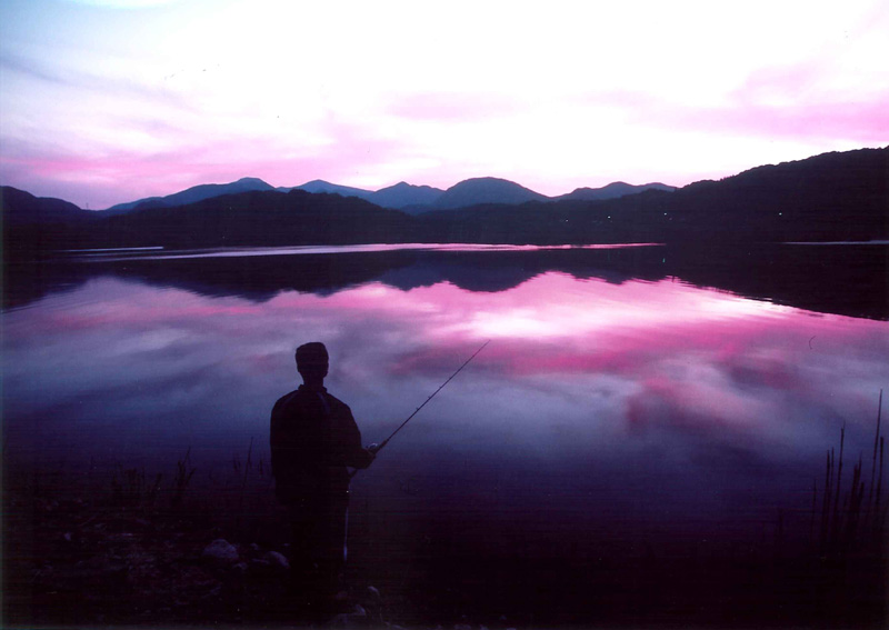ピンクと紫が混ざりあった空の色を反射している湖のほとりで釣りをしている人の写真