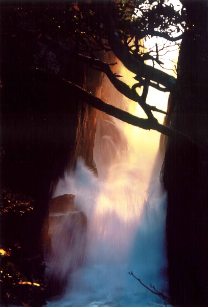 崖の間からオレンジ色の日差しが射し滝の水が流れている様子の写真
