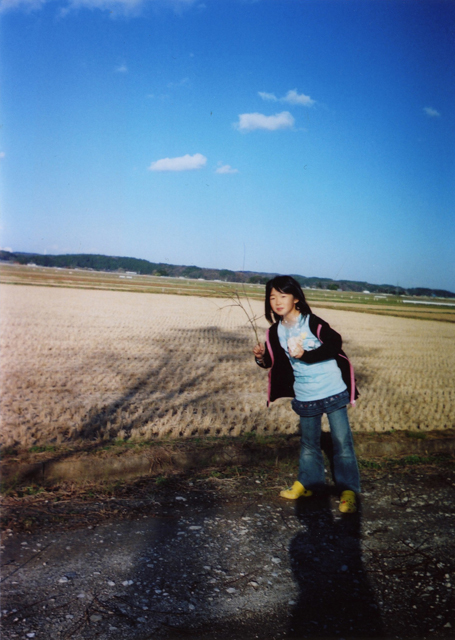 青空と田んぼを背景に子どもが木の枝を持ってポーズをとっている写真