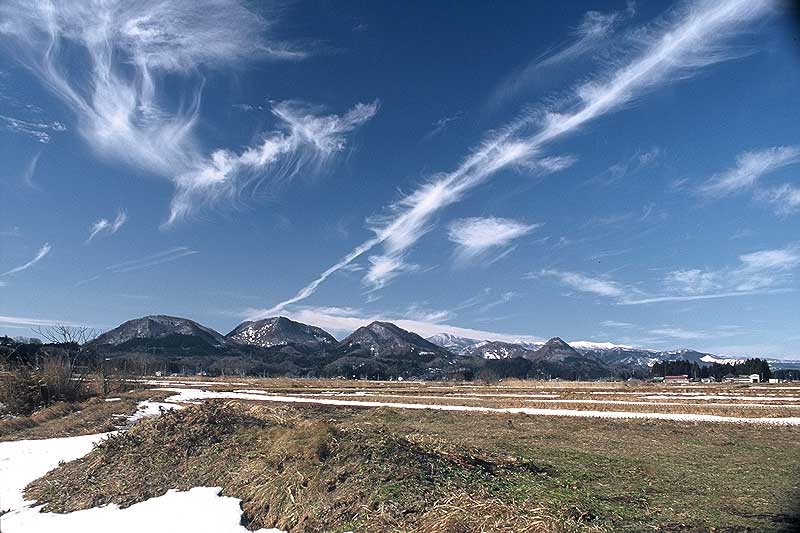 模様のような雲が出ている青い空と連なる山々と雪が残る野原を写した写真