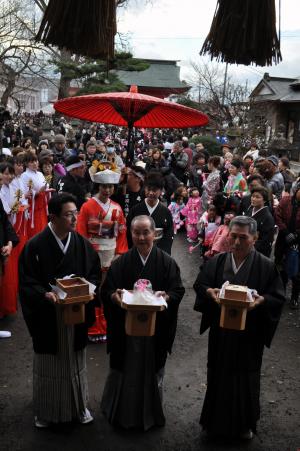 袴を着た三人の男性の後ろで赤い傘の下にいる二人の着物の女性の周りに大勢の人がいる写真