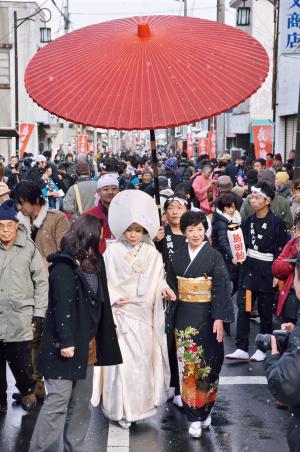 男性が差す赤い傘の下で白い着物を着た女性と黒い着物を着た女性が歩いている写真