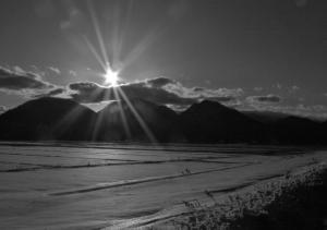 田んぼの先に見える山々と雲の上からのぞく太陽の白黒写真